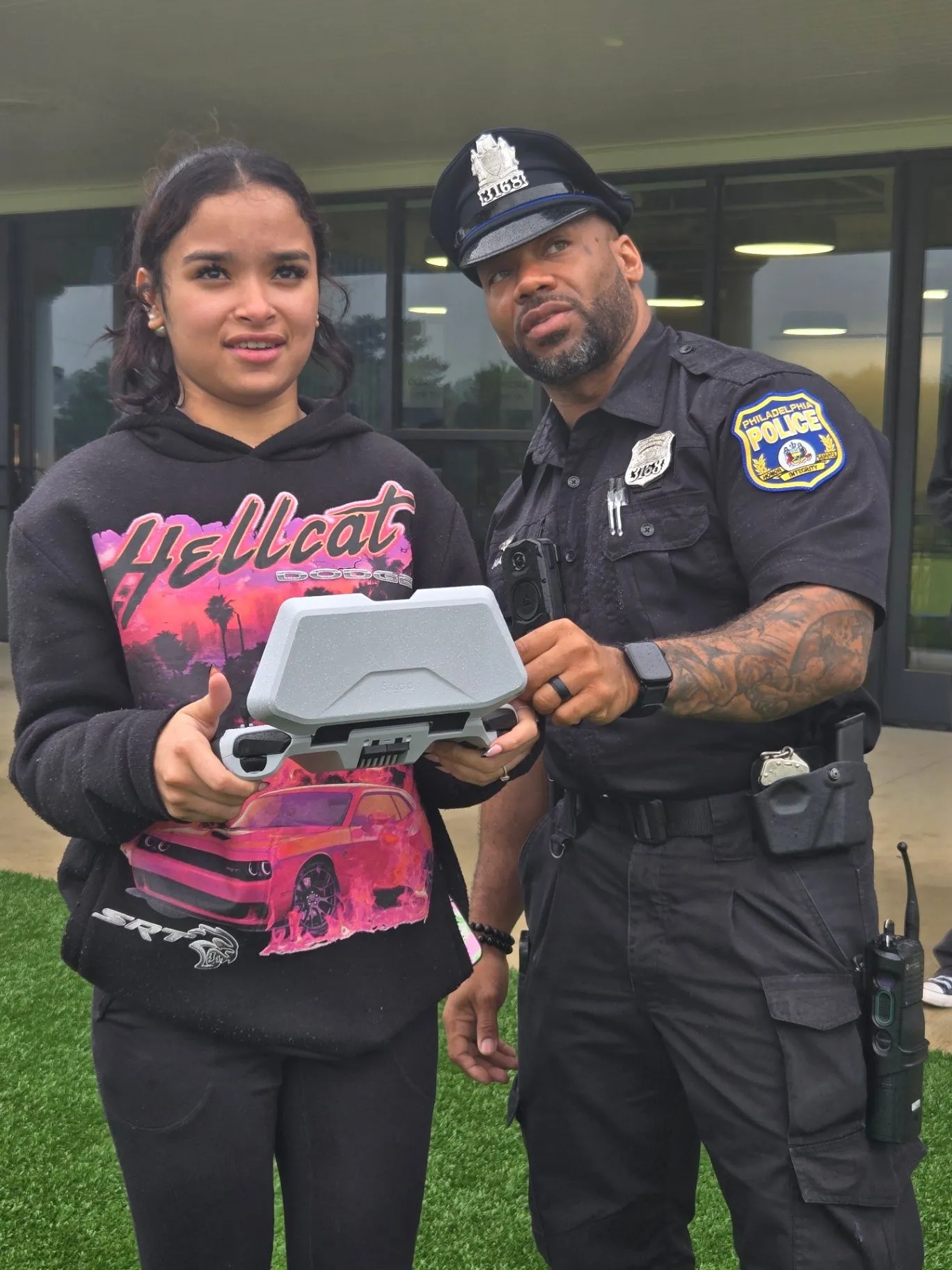 A student in a hoodie holding a drone with a smiling Philadelphia Police officer on a field