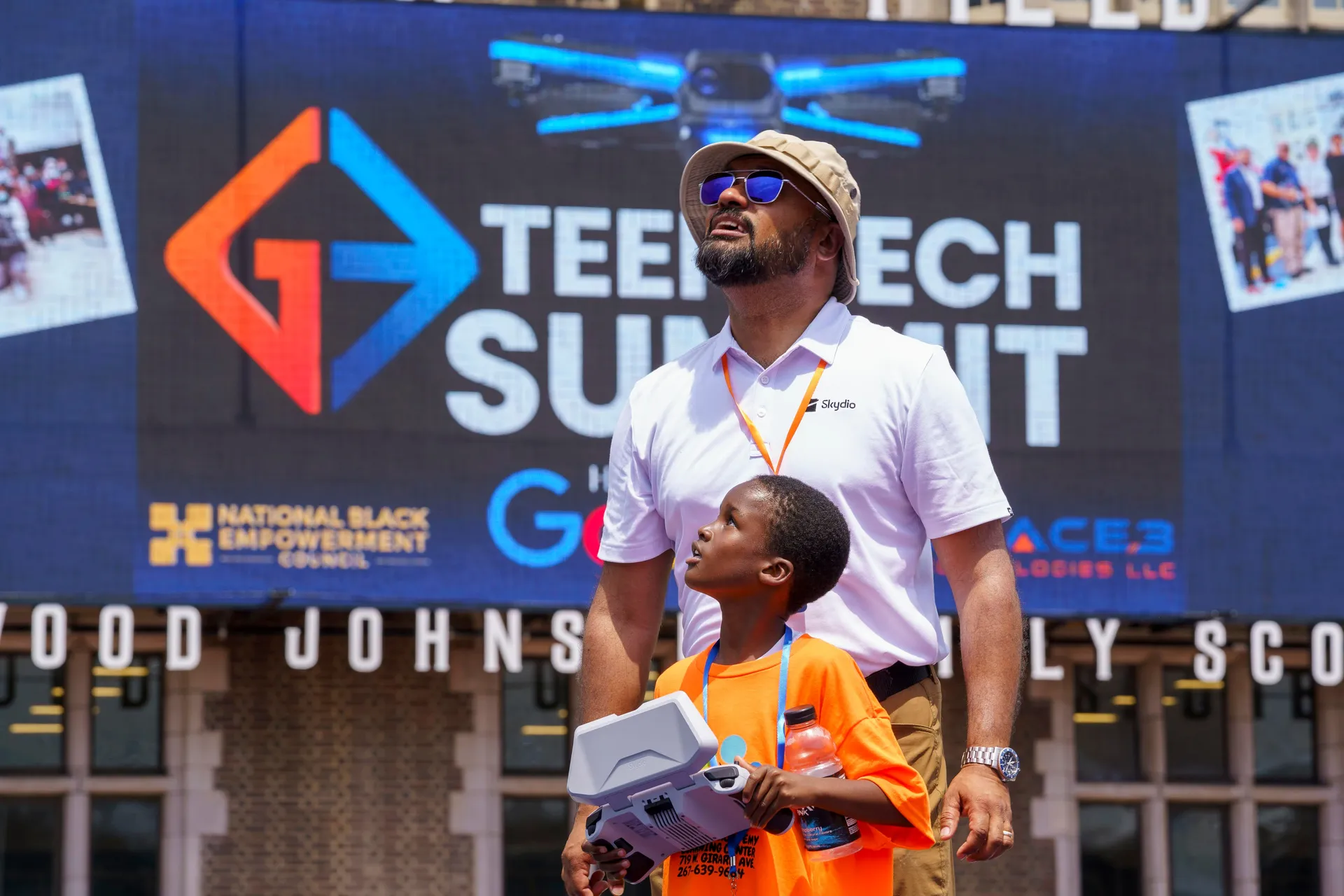 A boy holding a drone controller with a mentor in front of the Teen Tech Summit banner