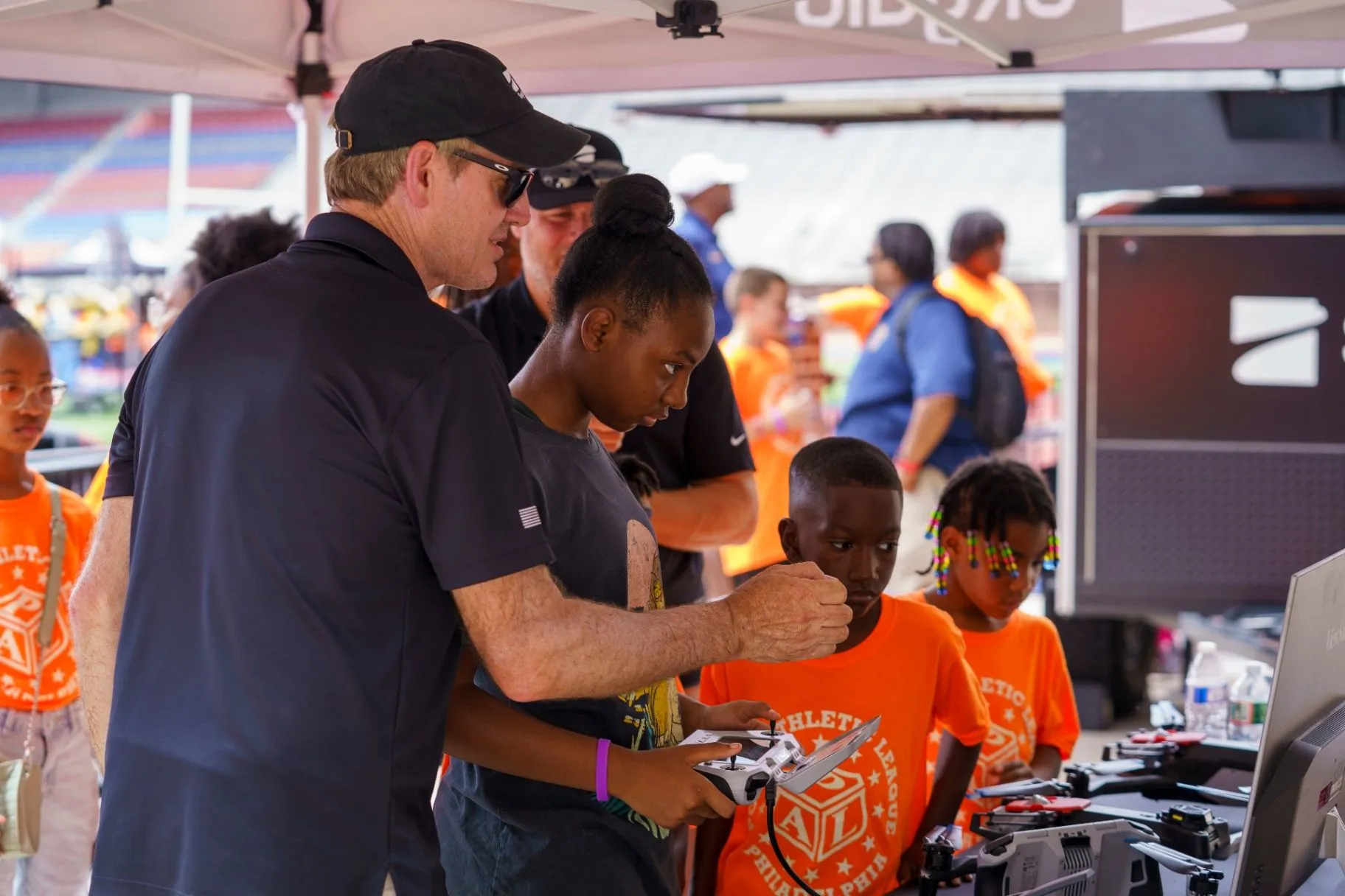 A mentor in a dark cap and sunglasses guiding a young girl holding a drone controller at a table of drones