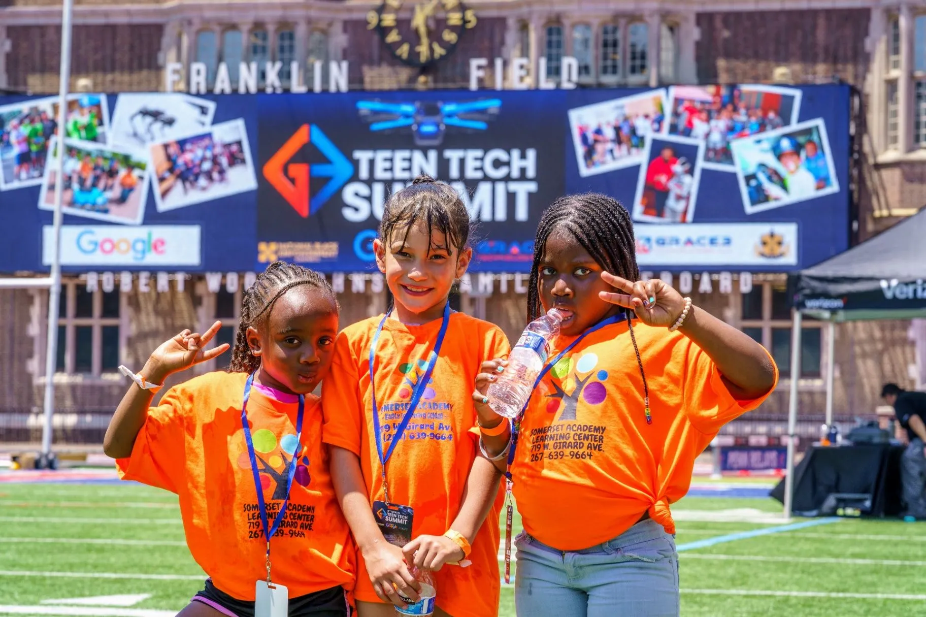 Three students in orange event shirts in front of the Teen Tech Summit banner at Franklin Field